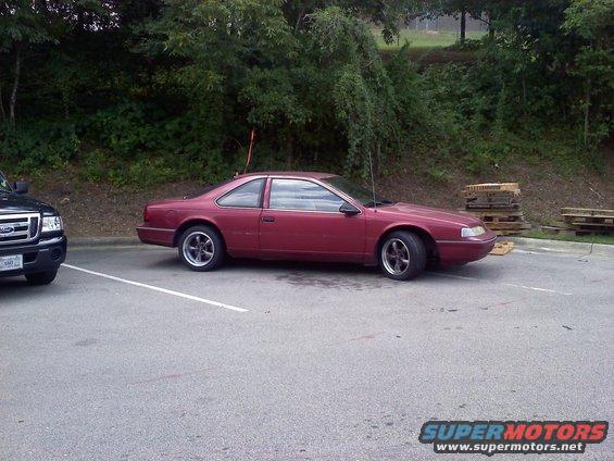 Maroon coupe parked next to a black pickup truck. Trees and pallets in background