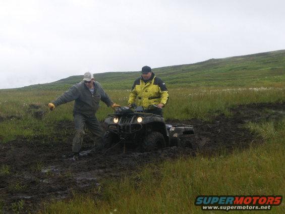 Man in gray pushes an ATV stuck in mud while another man in yellow jacket rides it