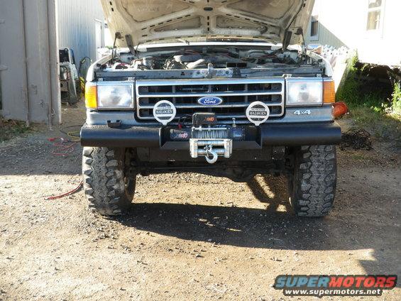 Front view of a Ford Bronco with hood open, showing engine and a winch mounted on the bumper. It has large off-road tires
