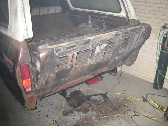 Rear of a 1979 Ford Bronco with significant rust and damage to the tailgate area. Some parts are removed, and welding equipment is nearby