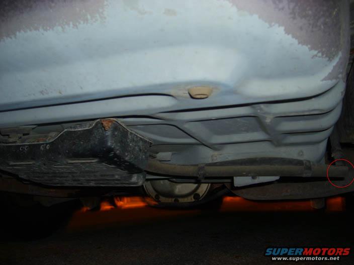 Underside of a vehicle showing a fuel tank, exhaust pipe, and differential. A red circle highlights a small component