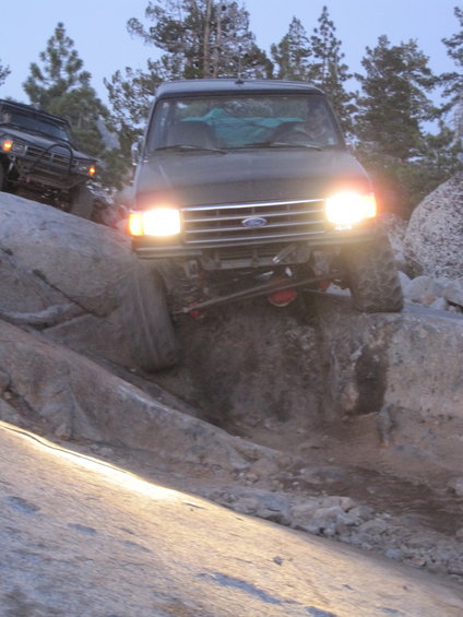 Dark 1989 Ford Bronco driving over rocks with headlights on