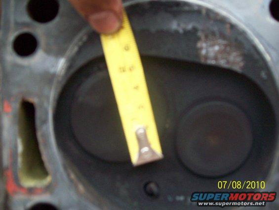 Close-up of an engine cylinder head with two valve openings. A yellow measuring tape is held over one opening
