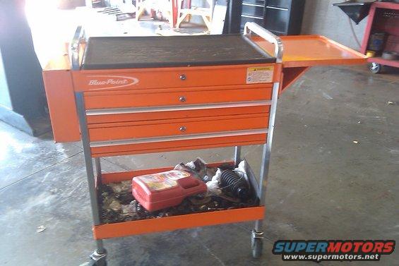 Orange tool cart with blue point logo, three drawers, and a pull-out side tray. Bottom shelf has a red container and some parts