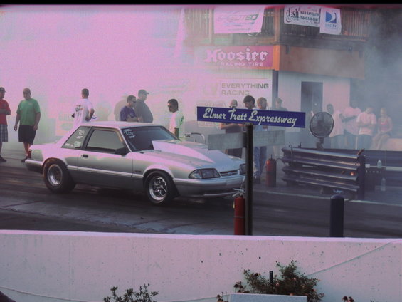 Silver coupe drag car with a large hood scoop, racing tires, and a roll cage visible through the window