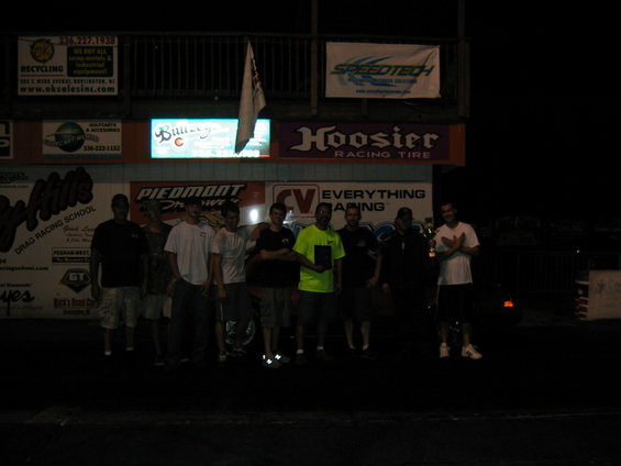 group of guys standing in front of drag strip signs at night, one holding a trophy