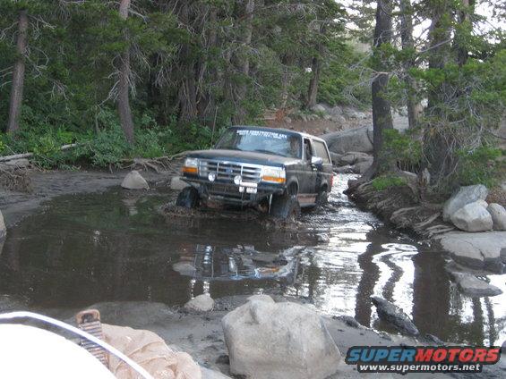 Black Ford Bronco driving through muddy water, front bumper modified with lights
