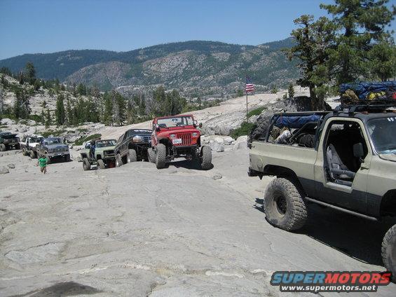Several off-road vehicles, including a red Jeep and a tan SUV, navigate a rocky, mountainous landscape