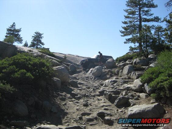 1986 Ford Bronco climbing a steep, rocky off-road trail with a person standing beside it