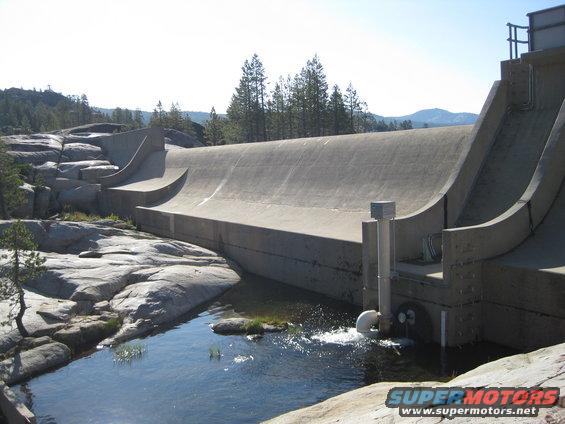 Concrete dam structure with water flowing out of a pipe into a pool below. Trees and rocky terrain surround it
