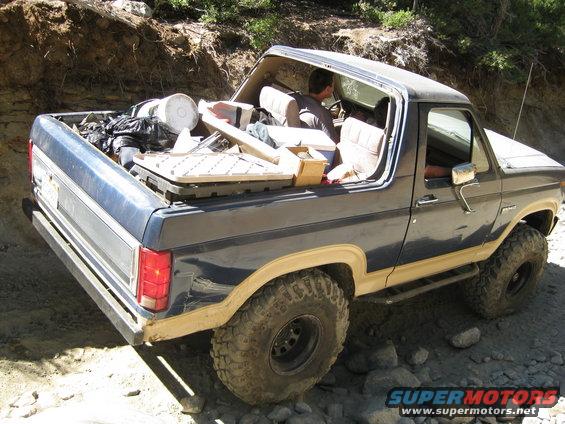 Dark blue Ford Bronco with tan trim, loaded with items in the bed and interior, driving on a rocky trail