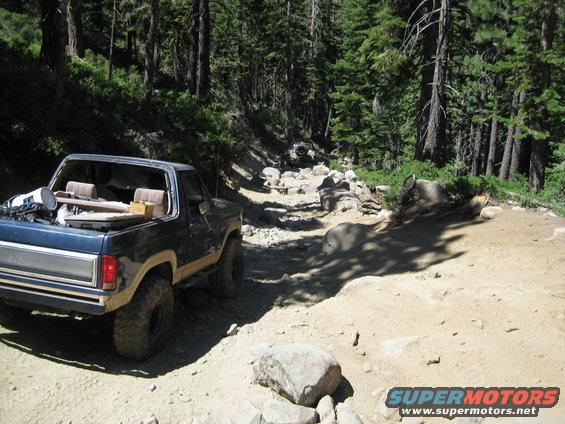 Blue Ford Bronco on a rough, rocky trail in a forest. Another vehicle is visible further up the trail