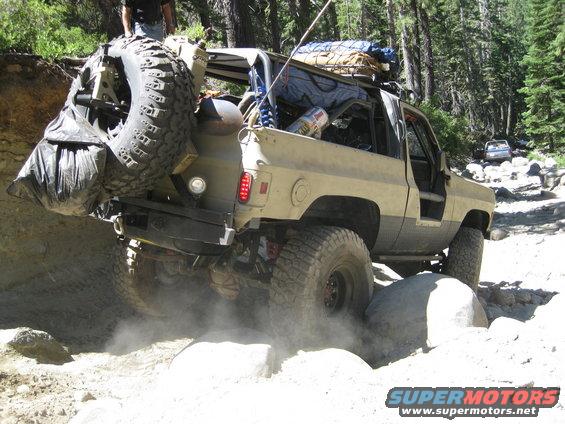 Modified off-road SUV with large spare tire on rear, navigating a rocky trail. Another vehicle is visible in the background