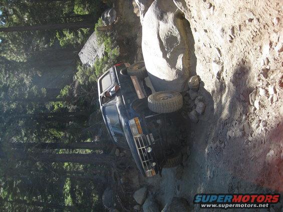 1986 Ford Bronco tilted on its side on a narrow, rocky trail with trees in the background