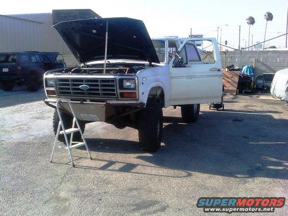 White Ford truck with hood open, front bumper, and driver door ajar. License plate visible