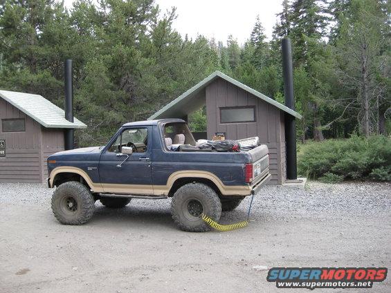 Blue Ford Bronco with large off-road tires parked next to a wooden outhouse