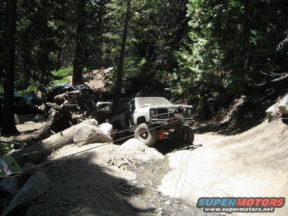 Off-road truck with lifted suspension and winch on a dirt trail, with another vehicle visible in the background among trees and rocks