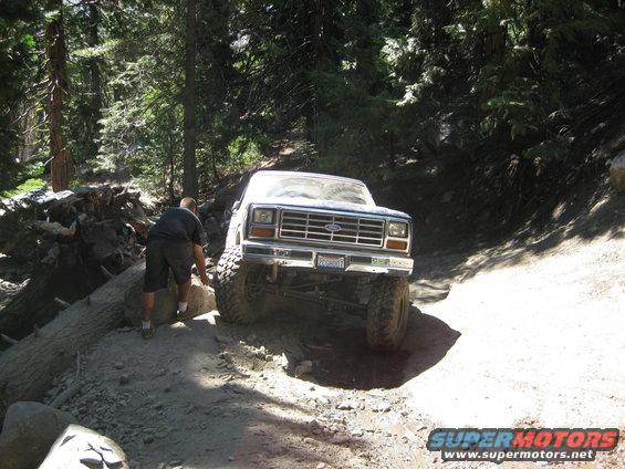 Ford Bronco with lifted suspension navigating a rocky, wooded trail. A person is outside the vehicle near the front tire