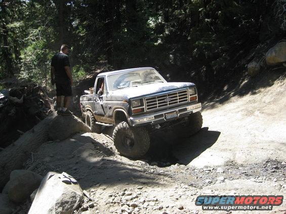 Ford pickup truck going off-road on a rocky trail. Driver is visible. Person stands on rocks nearby