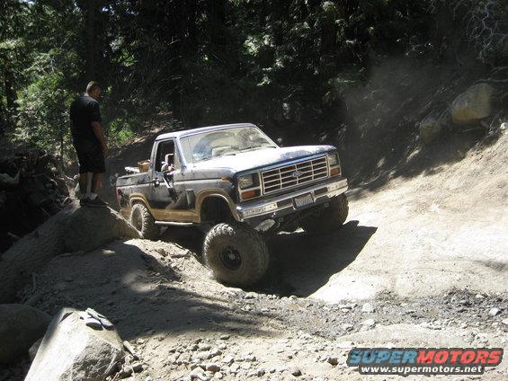 Dark blue Ford Bronco off-roading on a dirt trail, front-left tire on a rock
