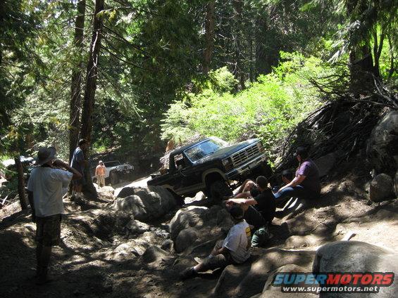 Black pickup truck navigating a rocky off-road trail with people watching