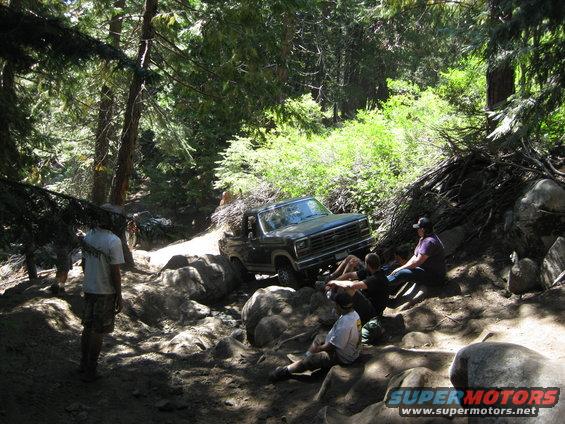 Dark green Ford Bronco navigating a boulder-strewn forest trail with people watching