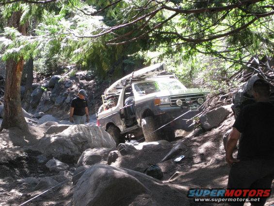 Ford Bronco navigating a steep, rocky off-road path with trees overhead. Two men are visible near the vehicle