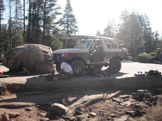 1991 Ford Bronco parked on rocks with people working on the front axle and a canoe on the roof rack