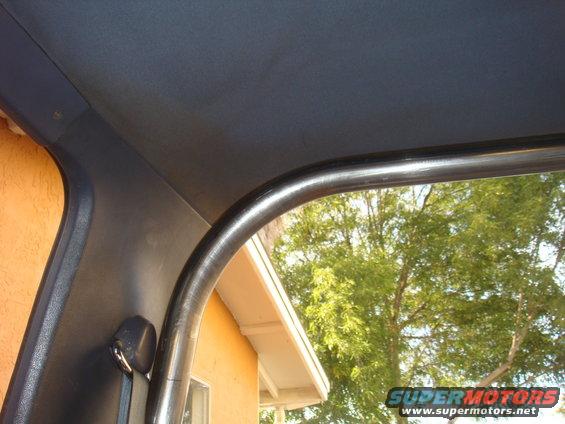 Interior view of a vehicle's roll cage with a seatbelt visible. Outside, trees and a house are seen