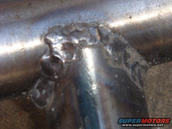 Close-up of a rough weld on shiny metal tubing. Supermotors.net logo visible