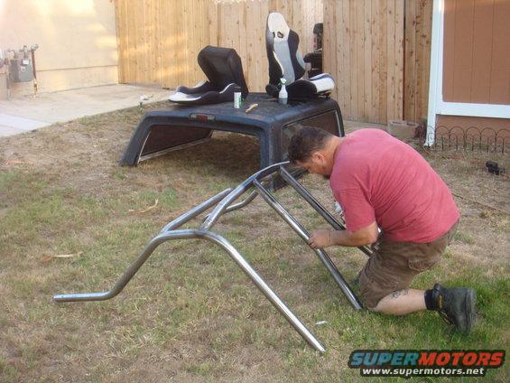 Man working on a metal roll cage in a backyard. Two racing seats are on top of a truck cap