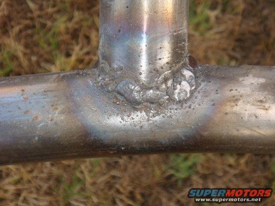 Welded metal tubing with rough welds and discoloration against a blurred grassy background