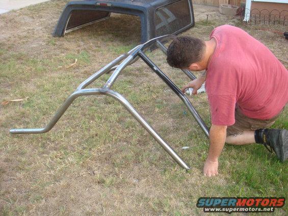 Man spray painting a metal roll cage on grass; truck bed cap in background