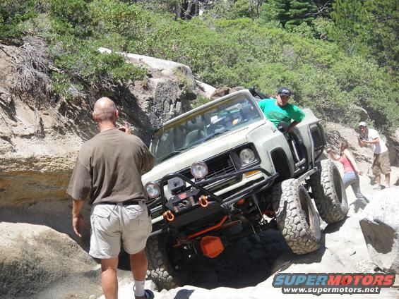 Modified off-road SUV with large tires climbing over rocks, people watching
