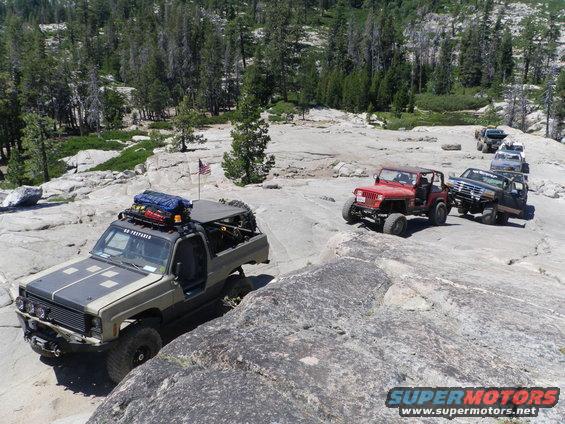 Several off-road vehicles navigate a rocky, wooded landscape. One vehicle in the foreground has a roof rack with gear