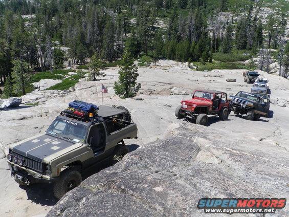 Several off-road vehicles driving on a rocky, wooded trail. An American flag is visible on one vehicle
