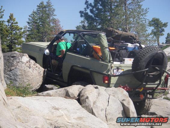 Olive green off-road SUV with spare tire on back, navigating large rocks. License plate reads 6APR540