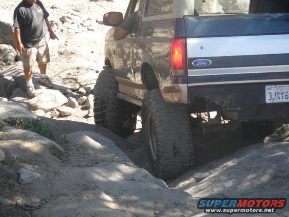 1991 Ford Bronco rear view, lifted with large tires, navigating rocky terrain. Ford badge visible
