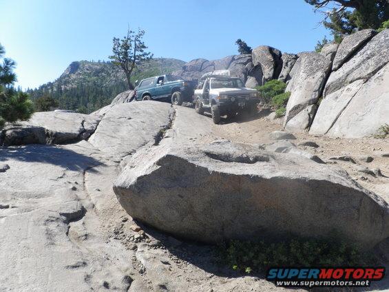 Green 1980s Ford Bronco and white off-road truck on a rocky mountain trail