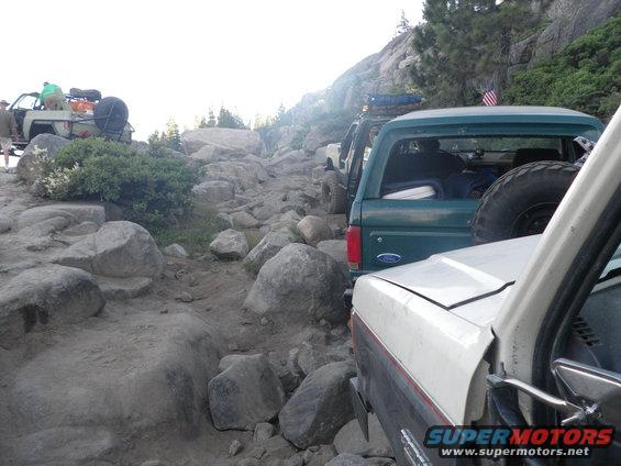 Line of off-road vehicles, including a 1991 Ford Bronco, navigating a rocky mountain trail