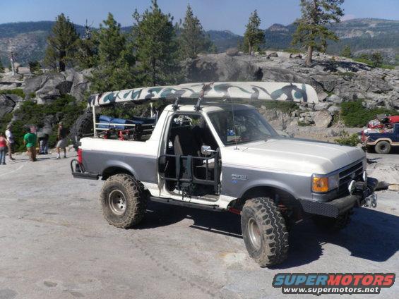 Two-tone Ford Bronco with doors removed, carrying a camo canoe on roof rack, parked on rocky terrain