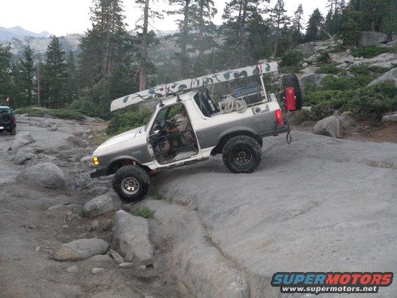 1991 Ford Bronco navigating rocky terrain, canoe secured to roof rack. Another SUV visible in background