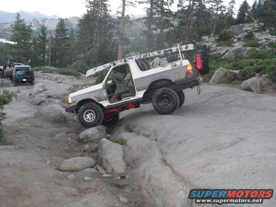 White and gray 1991 Ford Bronco with doors removed, navigating a rocky trail with a canoe on the roof rack