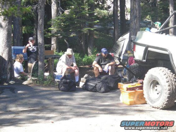 People are sitting around a campsite with a tent and a large off-road vehicle