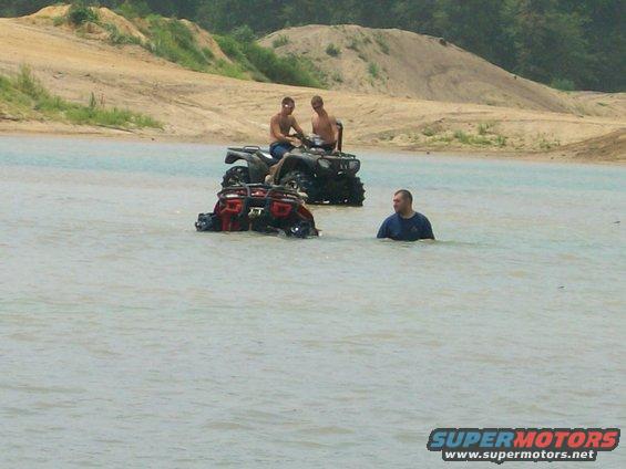 Three ATVs partially submerged in water, two with riders, one person standing in water