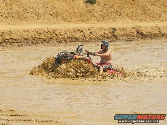 Person riding an ATV through muddy water, kicking up a large splash. Text overlay reads SUPERMOTORS