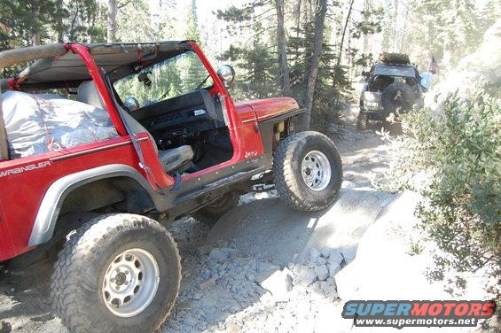 Red Jeep Wrangler with doors removed, driving on a rocky trail. Another Jeep is visible behind it