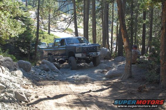 Modified dark gray pickup truck with large tires on a rocky trail, another vehicle visible behind it