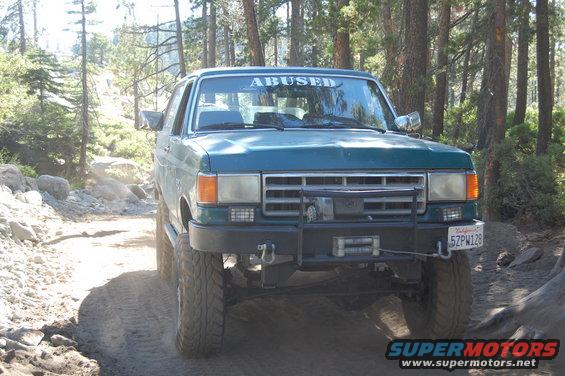 Teal Ford Bronco with "ABUSED" banner on windshield, lifted suspension, and winch