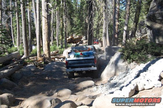Pickup truck navigating a boulder-strewn forest trail. Another vehicle is visible further up the path